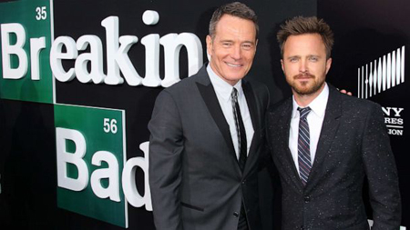 Bryan Cranston and Aaron Paul hug each in front of a Breaking Bad poster.